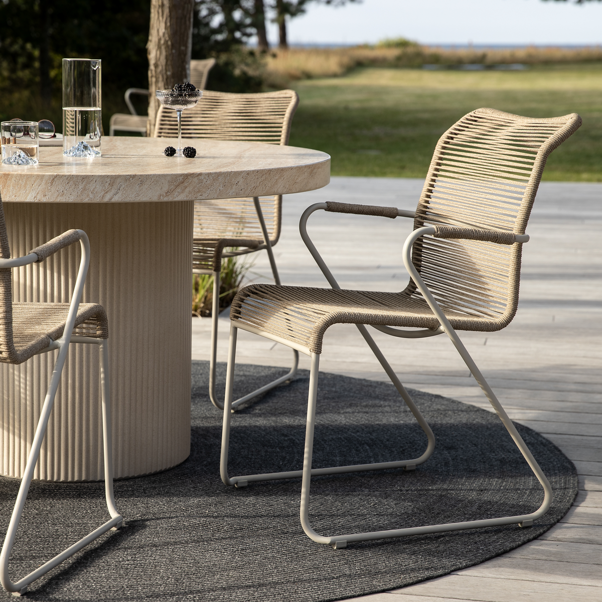 Udendørsscene med rundt stenbord og beige rebstole på et gråt rundt tæppe på en terrasse, med græsplæne og hav i baggrunden.