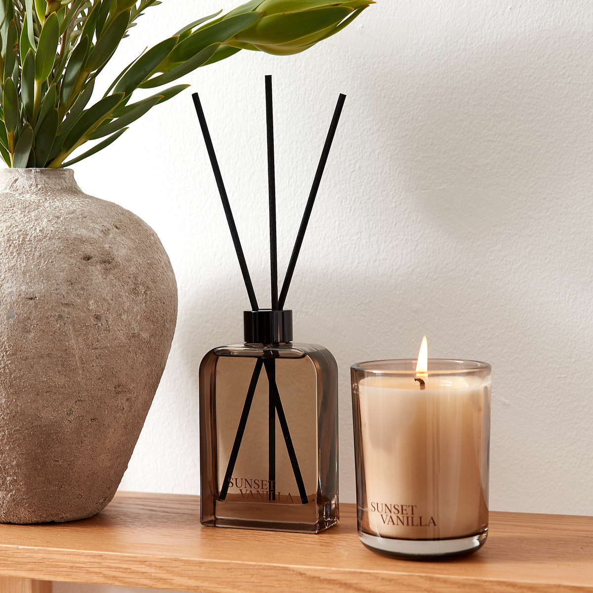 Duftspreder og tændt lys i brunt glas med Sunset Vanilla på en træhylde ved siden af en stenvase med grønne blade.