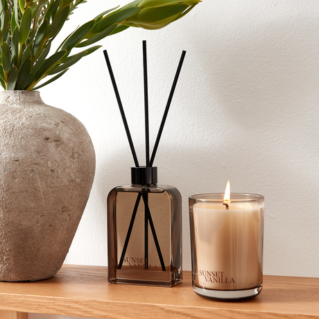 Duftspreder og tændt lys i brunt glas med Sunset Vanilla på en træhylde ved siden af en stenvase med grønne blade.