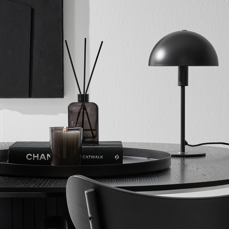 Sort bordmiljø med kuppelformet bordlampe, duftpinde og tændt lys på en bakke oven på bøgerne CHANEL og CATWALK.