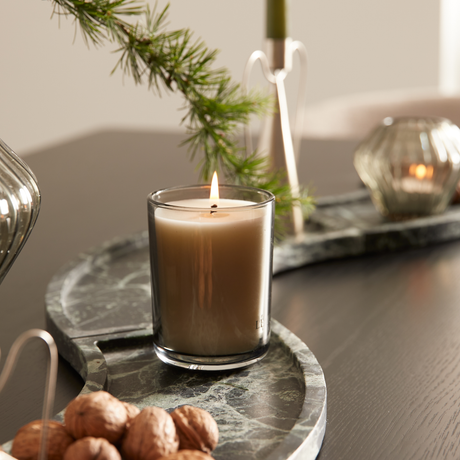 Tændt beige duftlys i glas på grøn marmorbakke med valnødder og gran, hyggelig borddekoration.