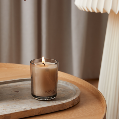Tændt duftlys i glas med duften Sunset Vanilla på en stenbakke på et træsidebord.