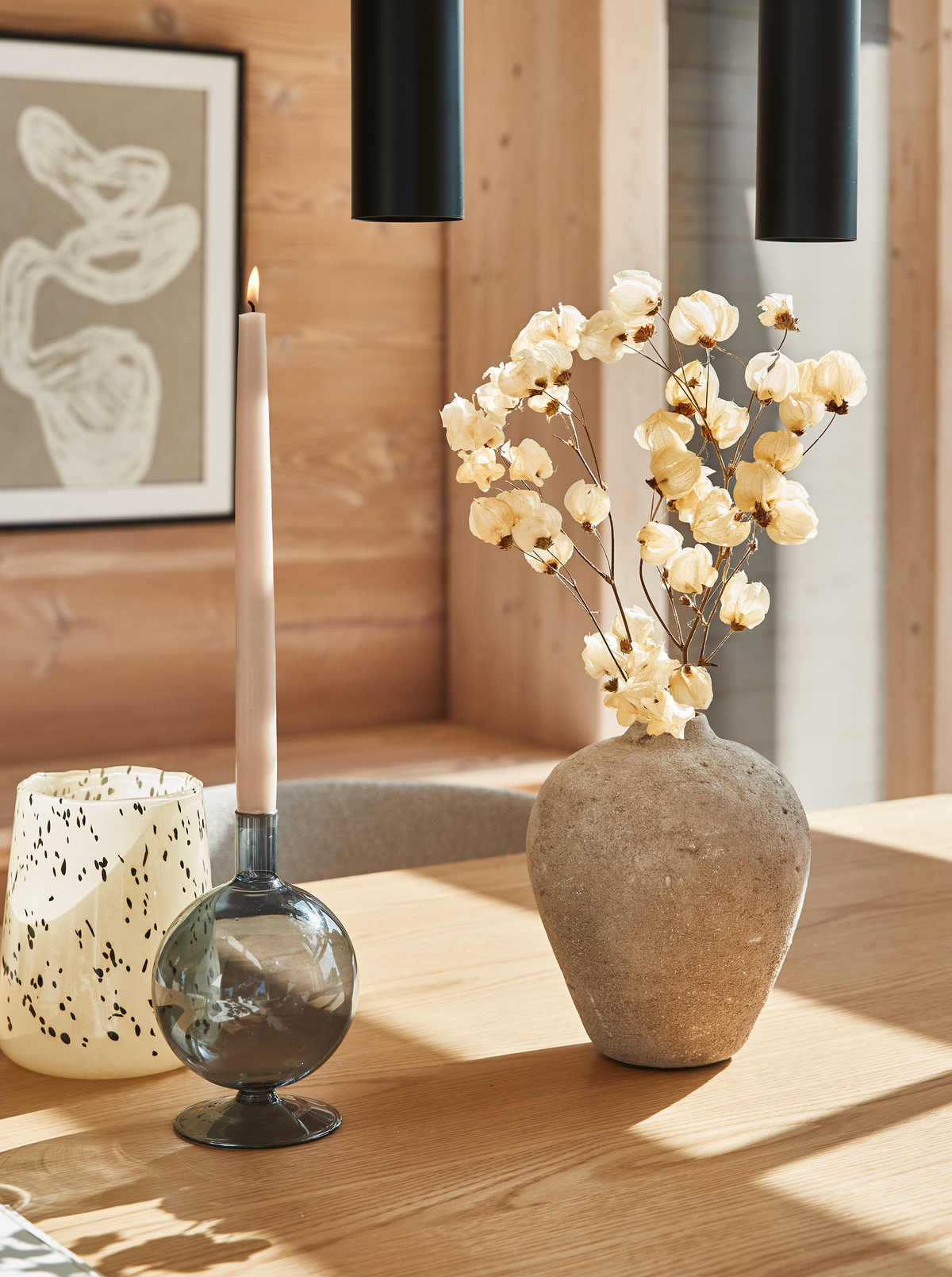 Stilleben på træbord med beige vase med tørrede blomster, et tændt slankt lys i røgfarvet lysestage og grafisk kunst i baggrunden i et solrigt, nordisk hjem.
