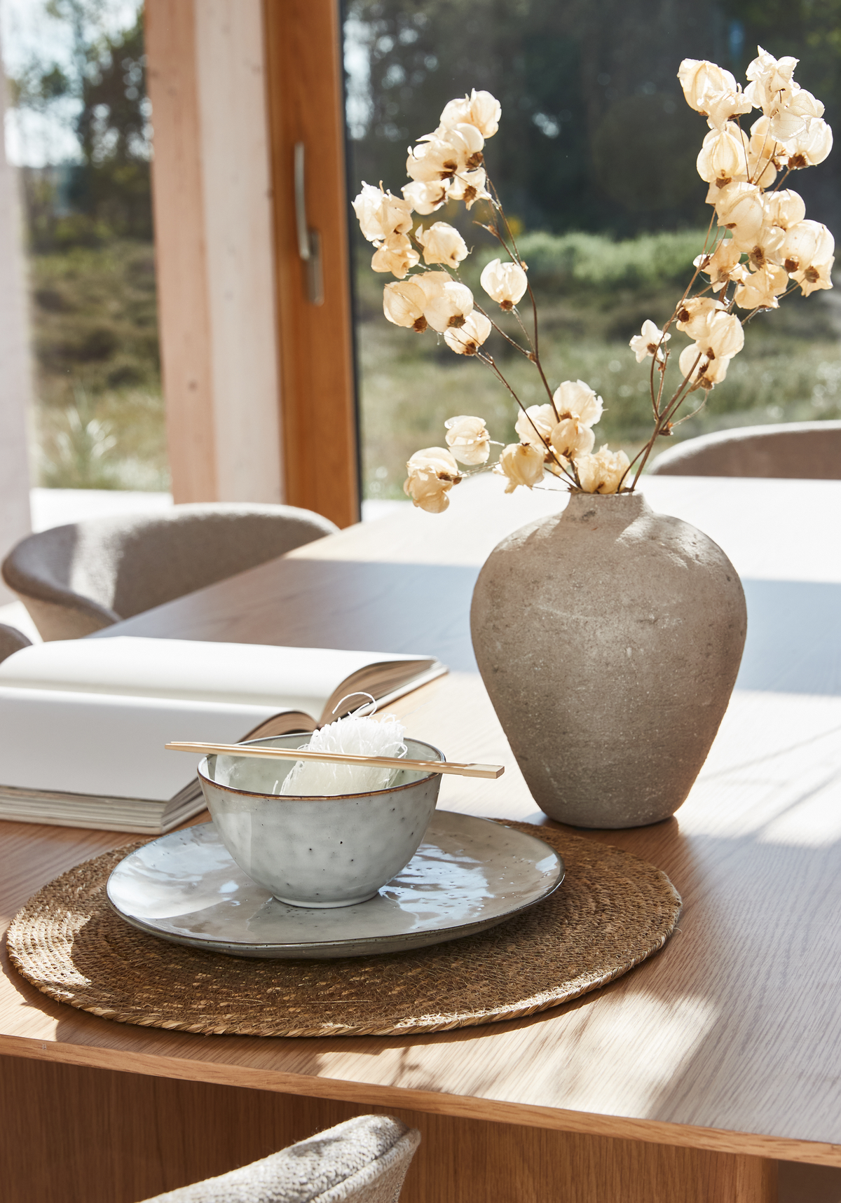 Stilleben på spisebord med keramikskål med spisepinde på tallerken, flettet dækkeserviet og stor vase med lyse tørrede blomster i sollys ved vinduet.