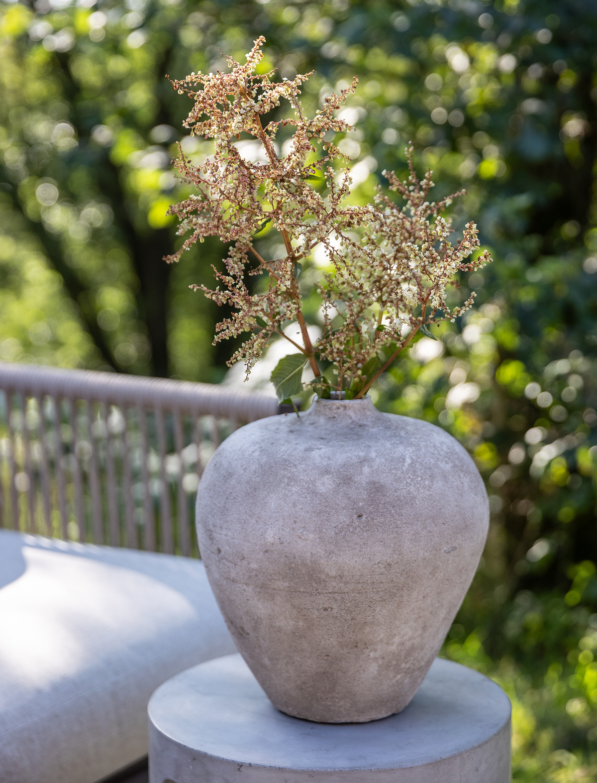 Betonlignende vase med gren med små blomster på rundt piedestalbord udendørs i sollys.