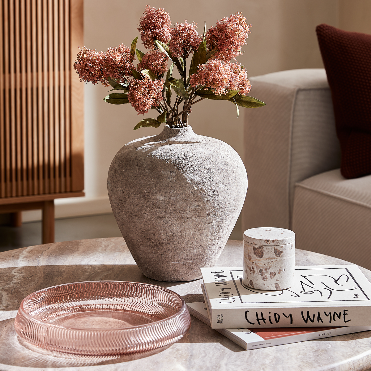Stenagtig vase med lyserøde blomster på et sofabord ved siden af et rosa glasfad, en terrazzo-krukke og bøger, ”CHIOY WAYNE” og ”Kinfolk”, i en lys stue.
