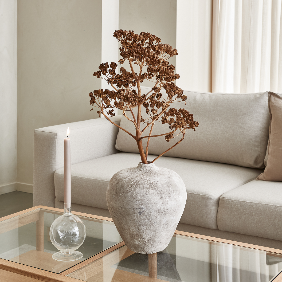 Beige stue med glasbord, rustik lerkrukke med tørret gren og tændt lys i glasstage foran en lys sofa.