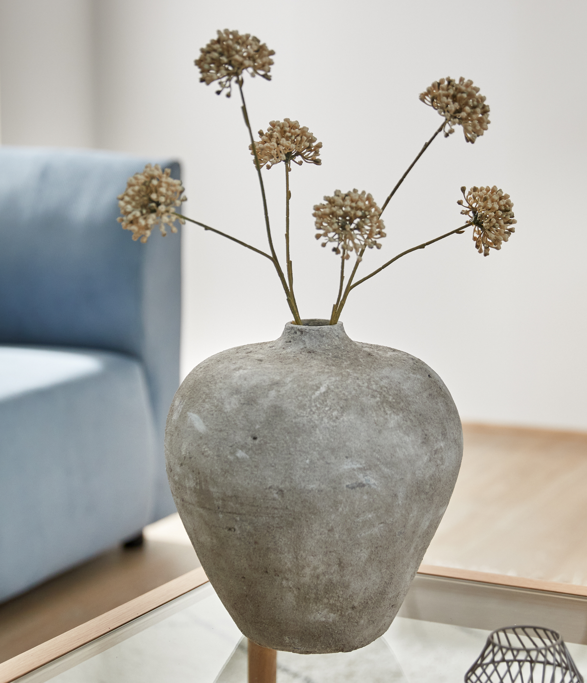 Stor grå betonlignende vase med tørrede blomsterstande på et glasbord i en stue.