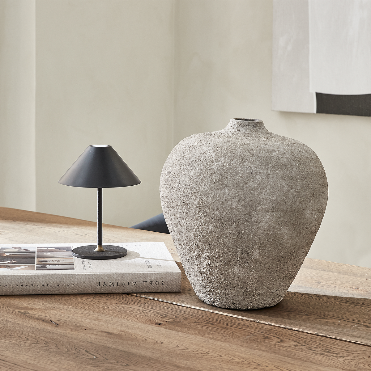 Rustik stenvase og lille sort bordlampe oven på bøger på et træbord i et lyst, minimalistisk rum.
