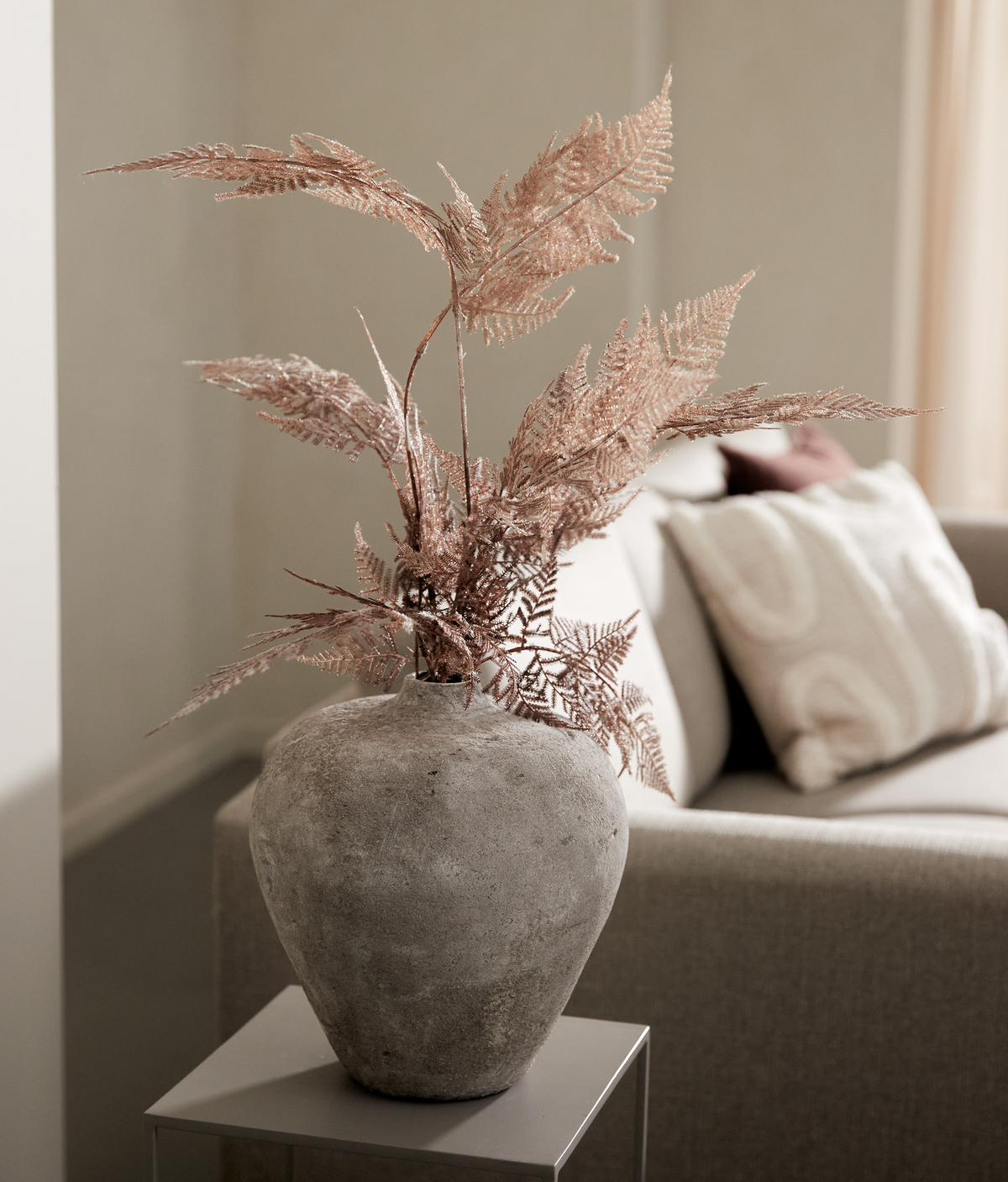 Rå betonvase med tørrede bregnegrene på et lille sidebord ved siden af en beige sofa med pude.