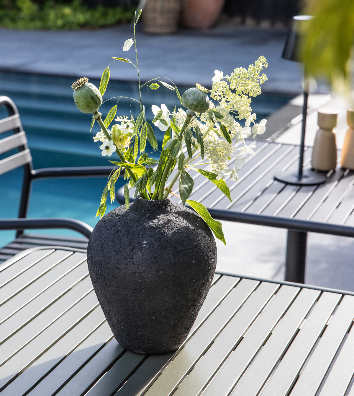 Mørk, struktureret vase med grønne kviste og hvide blomster på et gråt metalbord udendørs ved en pool.