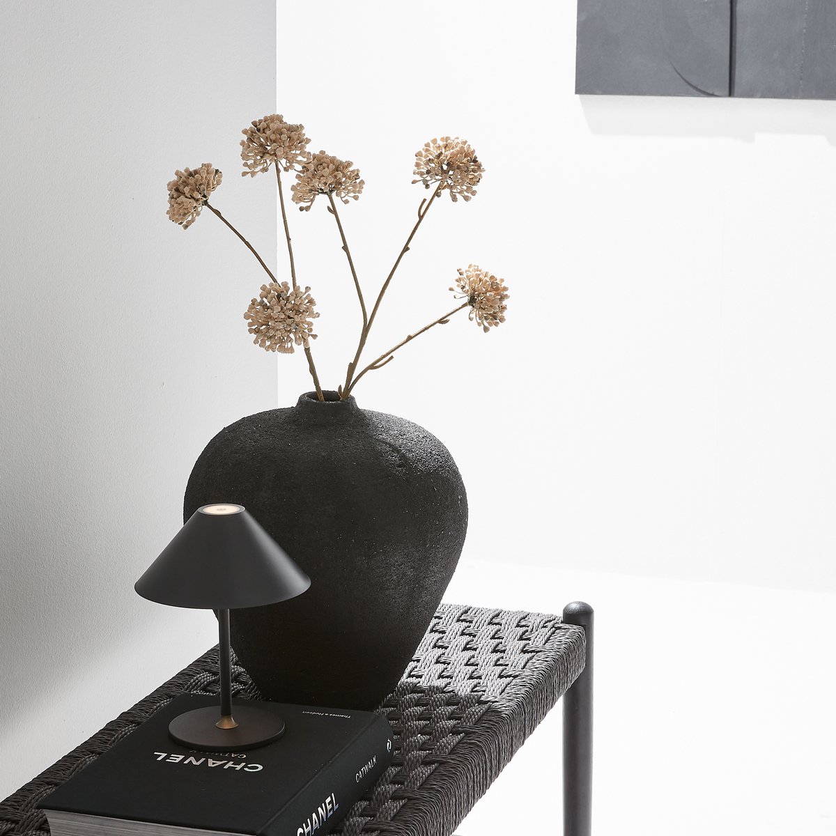 Sort, tekstureret vase med tørrede blomster ved siden af en lille sort bordlampe på en flettet bænk oven på bogen CHANEL i en lys, minimalistisk opsætning.