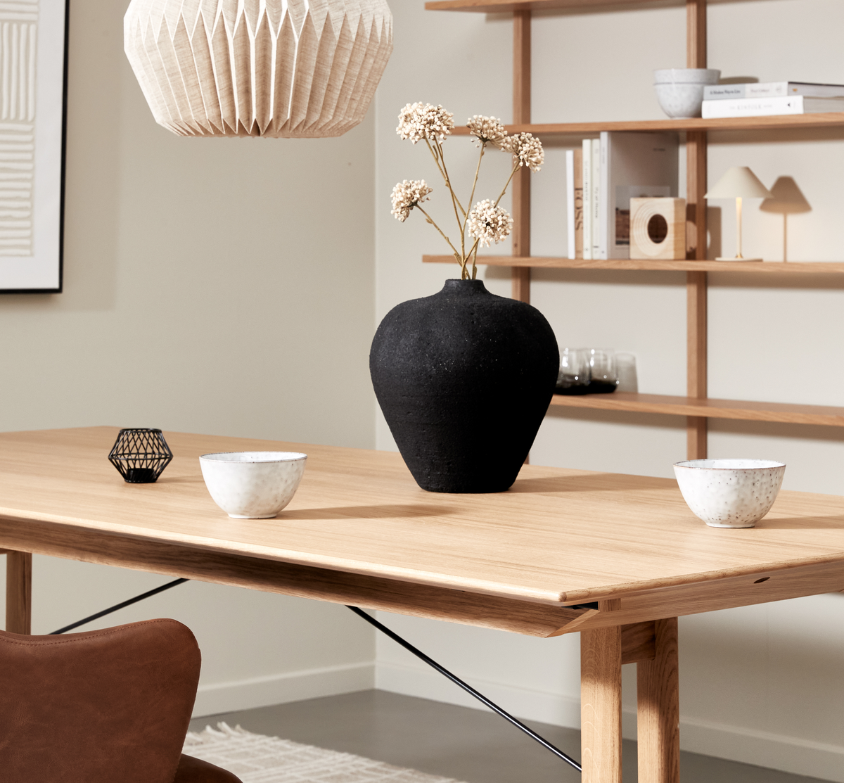 Lyst spisestue med egetræsbord, sort keramikvase med tørrede blomster, to hvide skåle og plisseret papirpendel; hylder med bøger og en lille lampe i baggrunden.