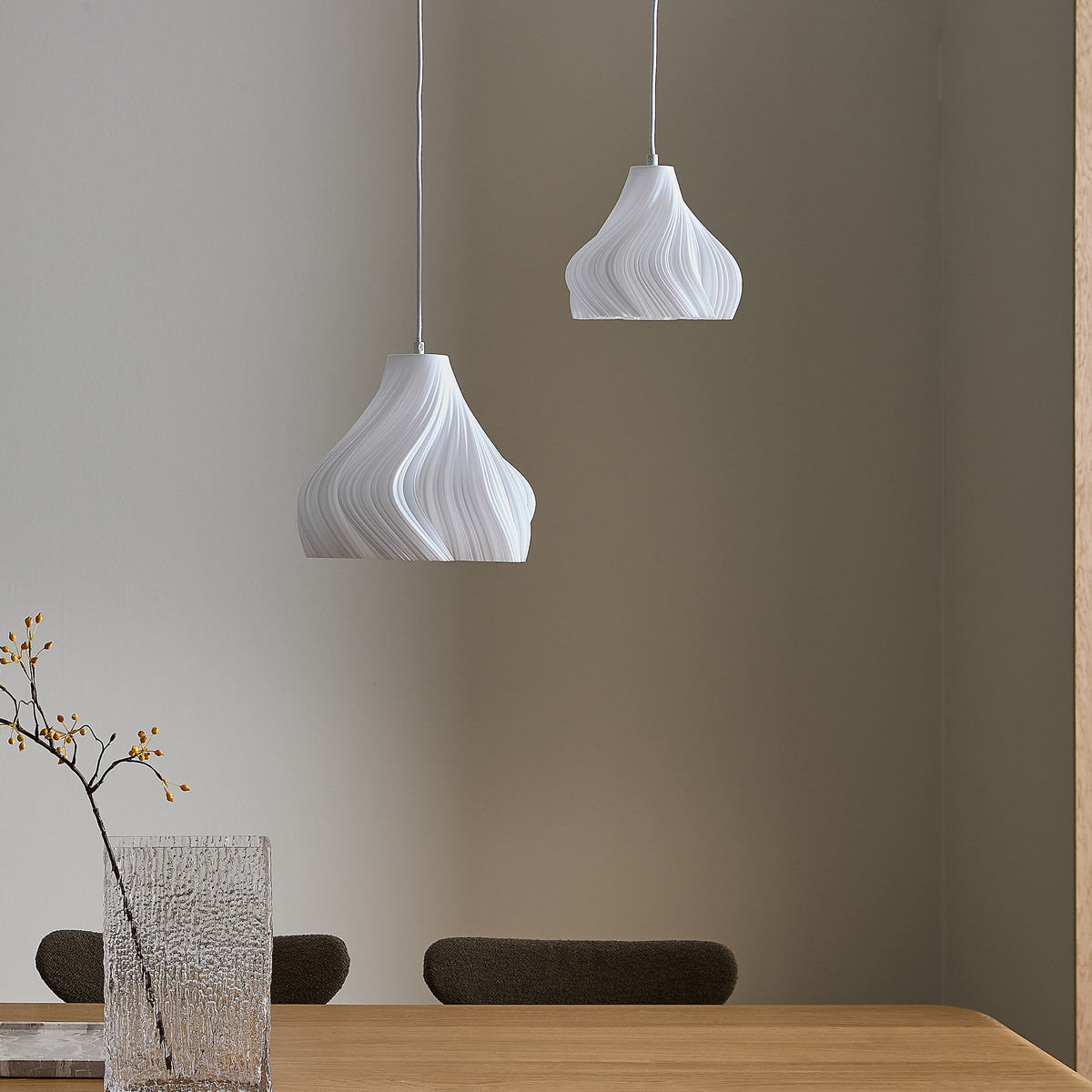 To hvide, skulpturelle pendellamper hænger over et træbord med en struktureret glasvase og en gren i en minimalistisk spisestue.
