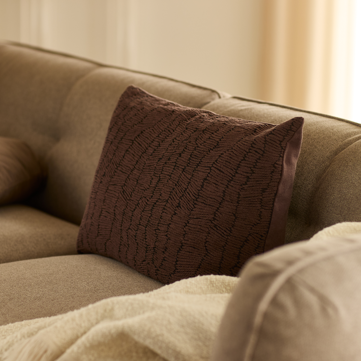 Brun, tekstureret pyntepude på beige sofa med lys plaid i blødt, varmt lys.
