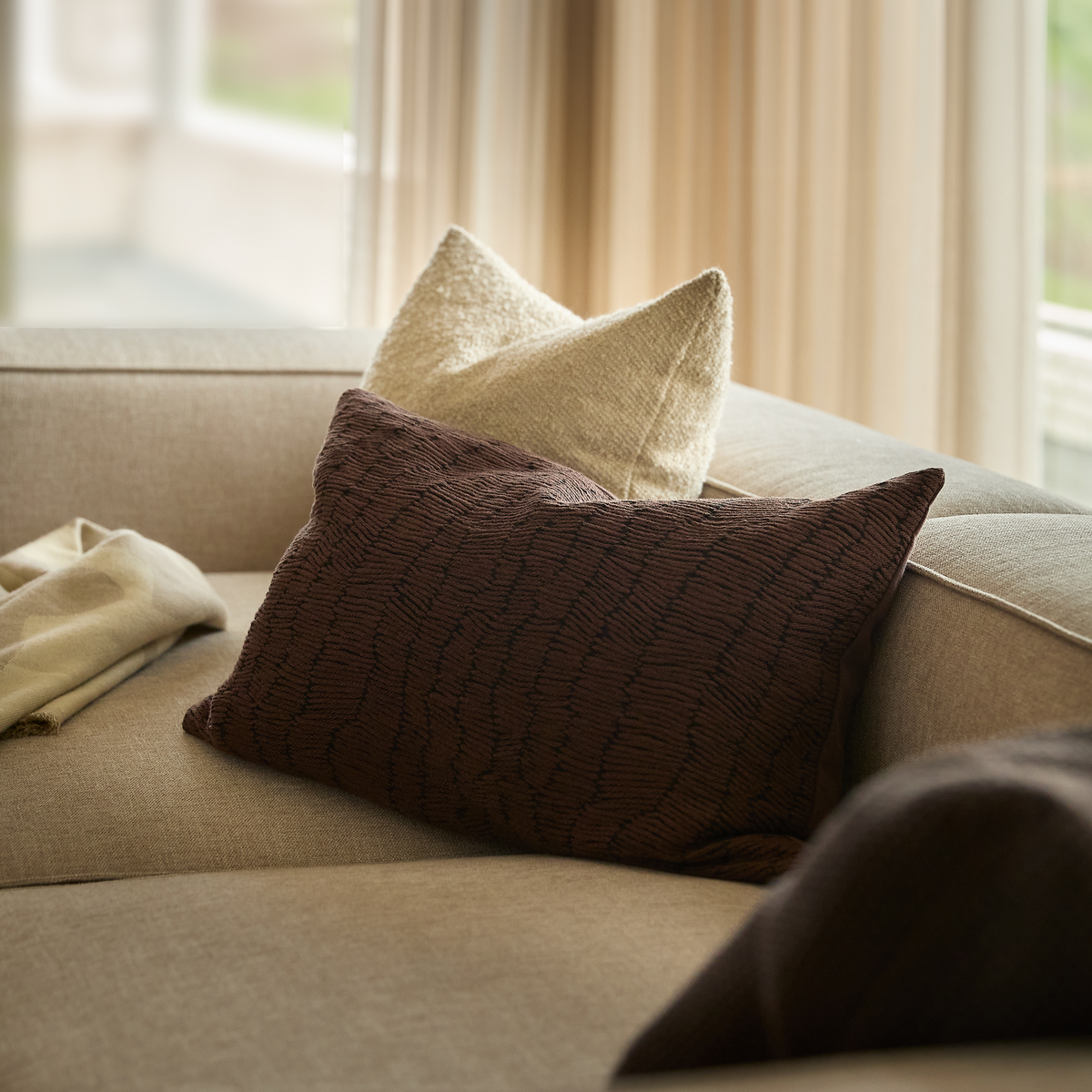 Nærbillede af beige sofa med en mørkebrun, tekstureret aflang pude og en cremefarvet pude, med bløde gardiner i baggrunden.