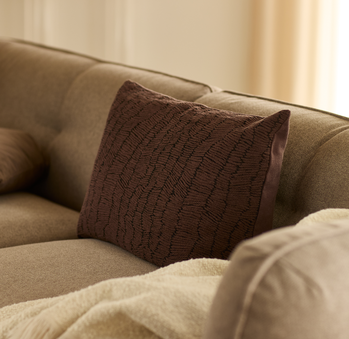 Brun struktureret pyntepude på beige sofa med lys plaid i et varmt stue-miljø.