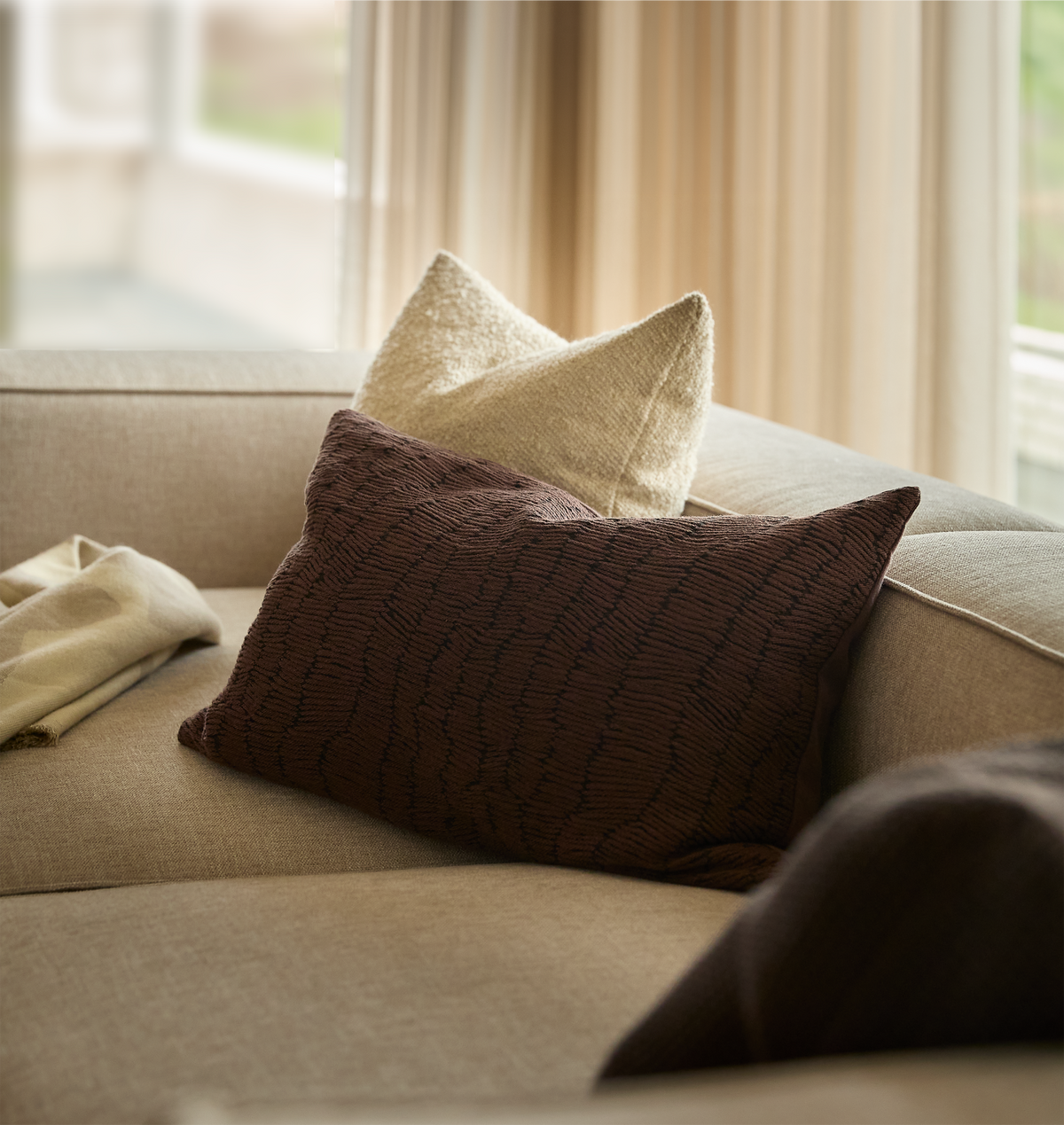 Beige sofa med brun rektangulær pude og en cremefarvet pude i blødt dagslys foran gardiner.