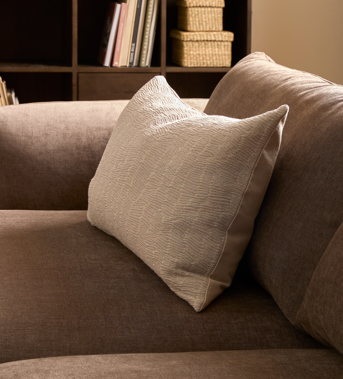 Beige, struktureret pyntepude på brun sofa i en stue med reol i baggrunden.