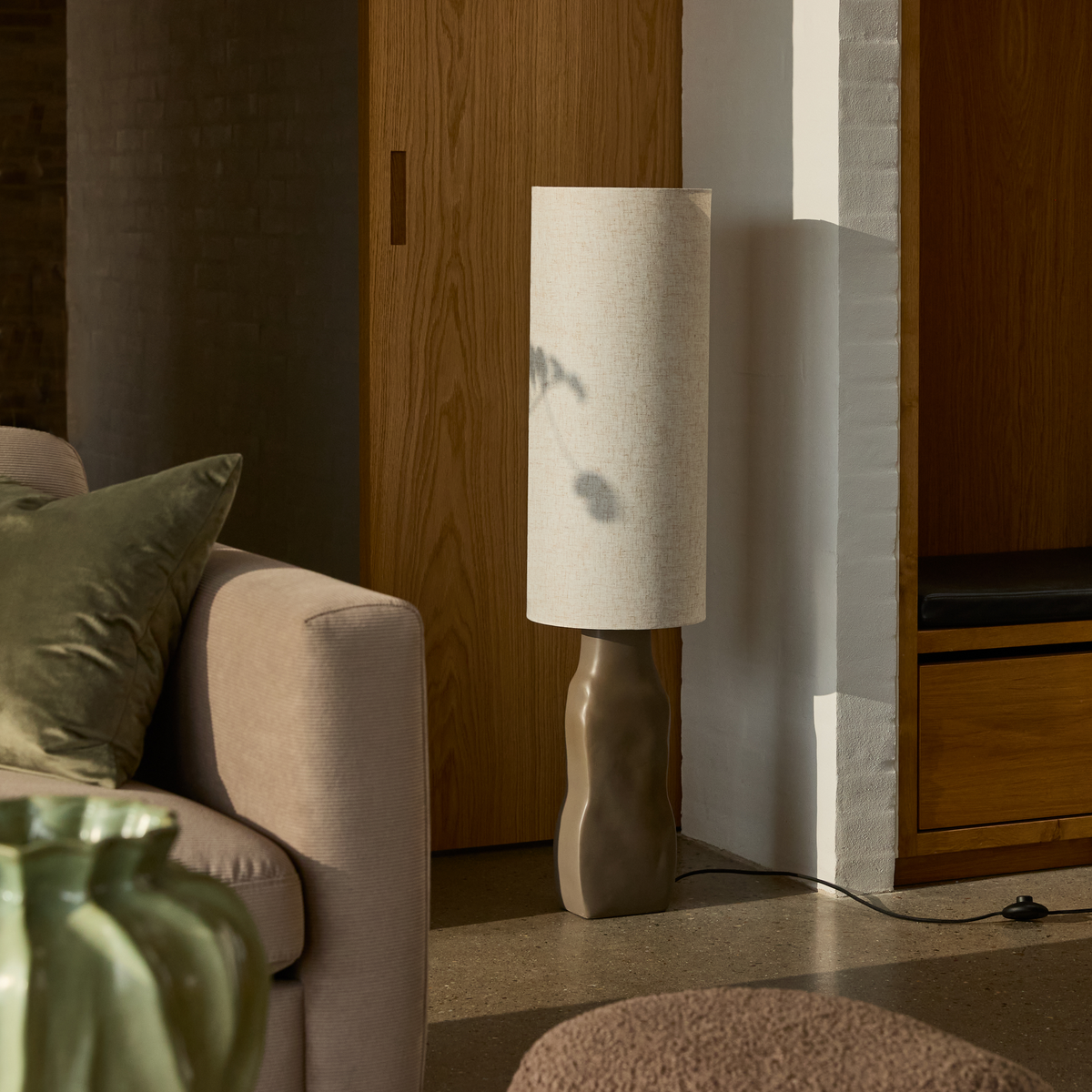Skulpturel gulvlampe med høj tekstilskærm ved siden af beige sofa og træskab i en hyggelig stue.