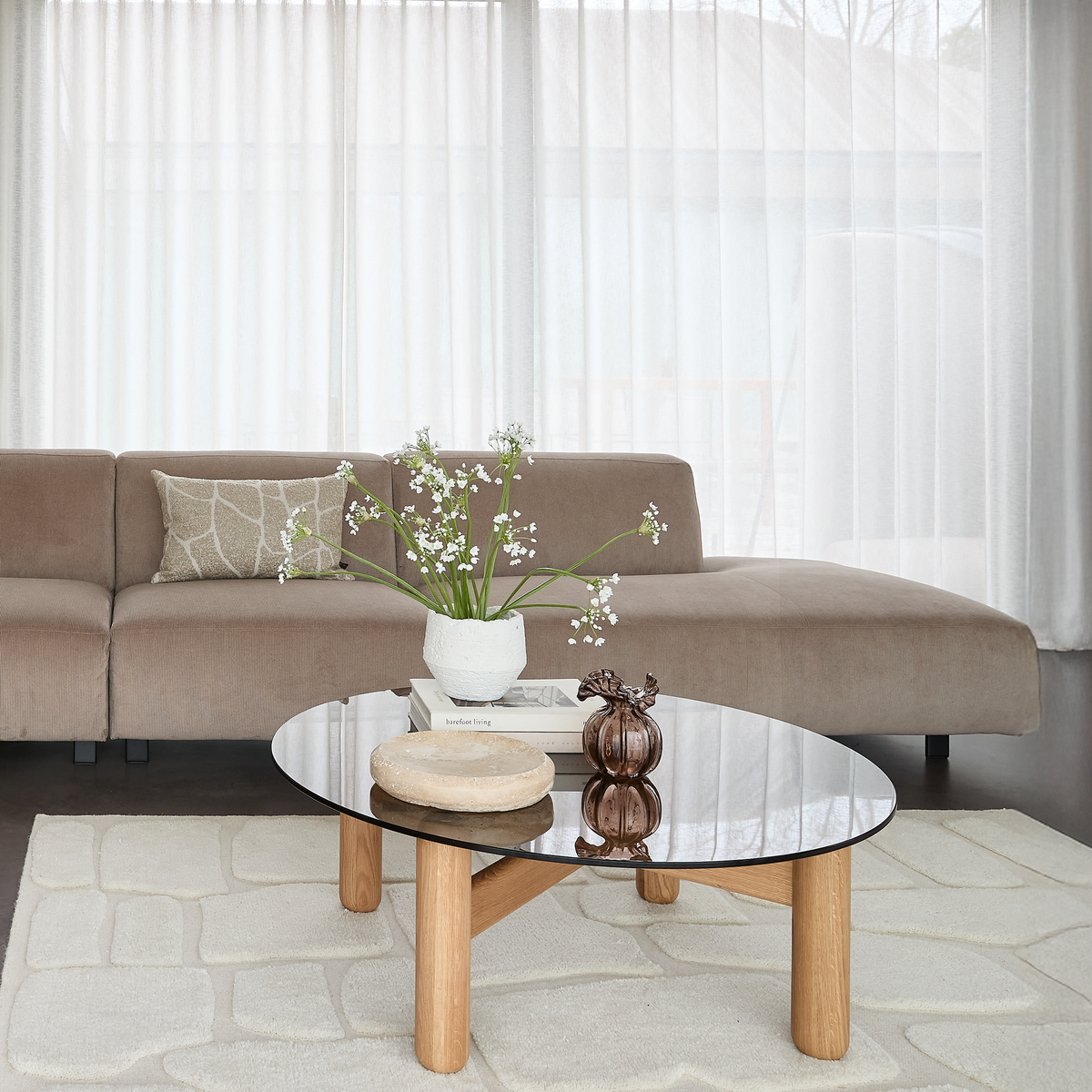 Lyst stueinteriør med beige modulsofa, glasbord med træben, vase med hvide blomster oven på bogen ”Herbarium Living”, skulptur og stenskål, lette gardiner og lyst tæppe.