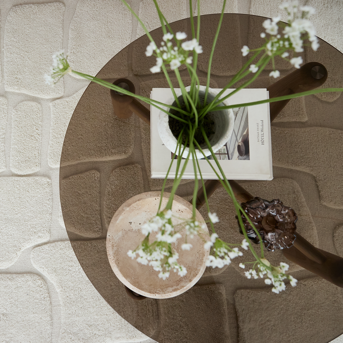 Set oppefra af et rundt sofabord i glas med en lille potteplante med hvide blomster, en bog, et stenfad og et dekorativt objekt på, over et beige tekstureret tæppe.
