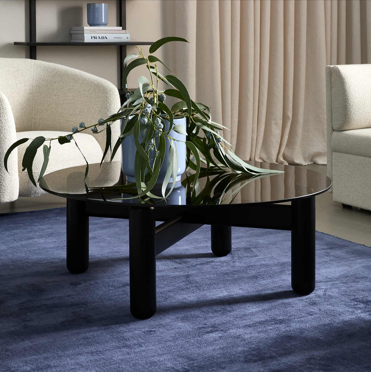 Sort rundt sofabord med blank top på blå tæppe, en blå vase med grønne grene ovenpå; beige gardiner og lyse lænestole i baggrunden, bog med titlen PRADA på hylden.