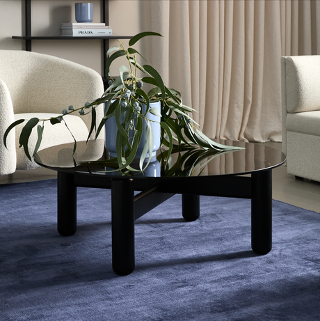 Sort rundt sofabord med blank top på blå tæppe, en blå vase med grønne grene ovenpå; beige gardiner og lyse lænestole i baggrunden, bog med titlen PRADA på hylden.
