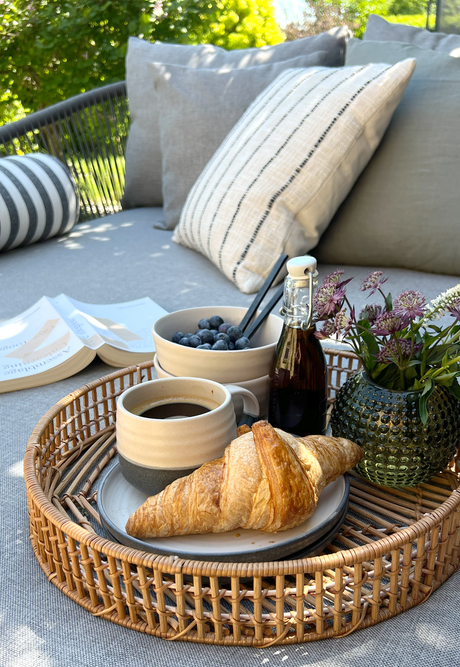 Morgenmadsbakke med kaffe, croissant, skål med blåbær, lille glasflaske og grøn vase med blomster på en udendørs sofa med grå og stribede puder i sollys.
