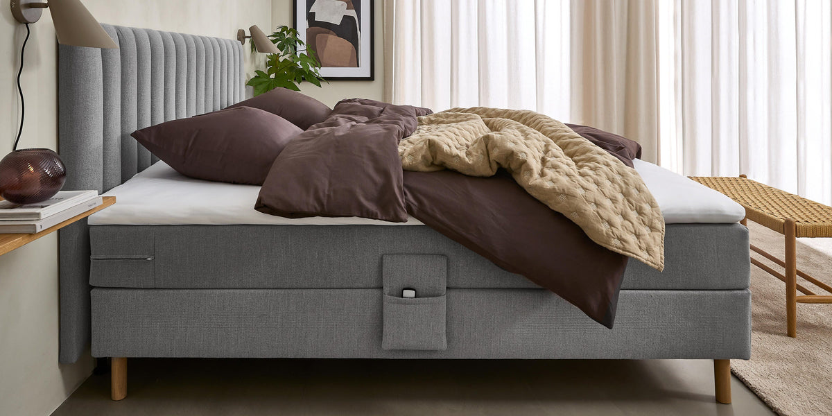 Lyst soveværelse med grå polstret seng, brunt sengetøj og beige quilt; lyse gardiner, væglamper, lille hylde med bøger og glaslampe, plante og kunst.