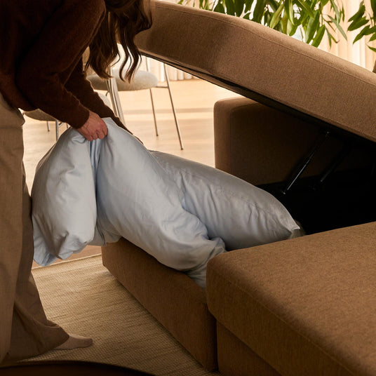 Person lægger en lyseblå dyne i opbevaringsrummet under en brun sofa i en lys stue med planter.