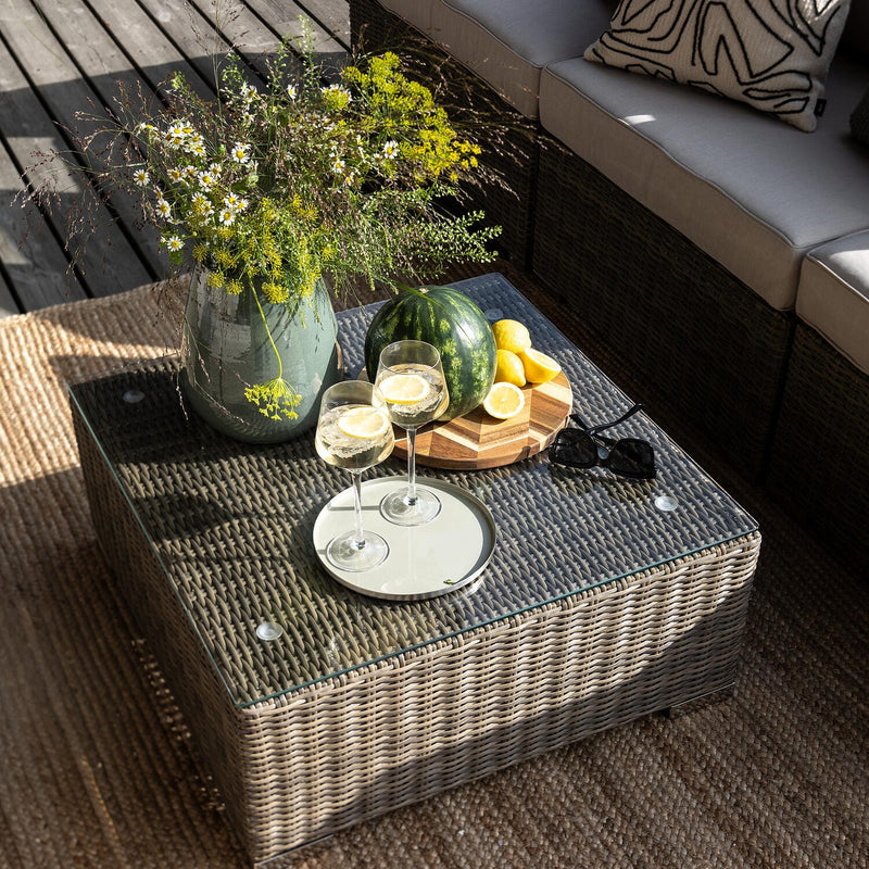 Udendørs sofahjørne med fletbord med glasplade, vase med vilde blomster, skærebræt med hel vandmelon og citroner, to glas med citronskiver og solbriller i sollys.