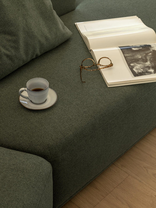 Grøn sofa med opslået bog, læsebriller og espressokop på underkop.