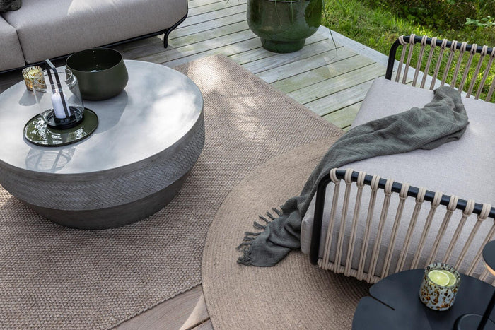 Udendørs loungemiljø på træterrasse med rundt beton-sofabord med lyslygte og skål, loungestol med snordetaljer og gråt plaid, neutrale tæpper og grøn plantekrukke i dagslys.