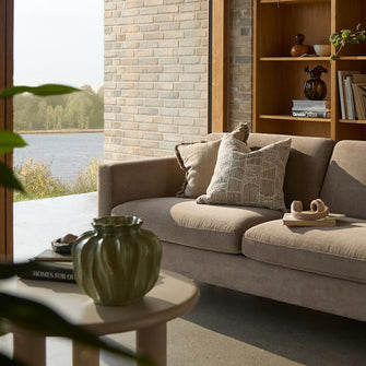 Lyst stue med beige sofa foran en træreol, sofabord med grøn vase og bogen Homes for Our Time, udsigt til en sø gennem stort vindue.