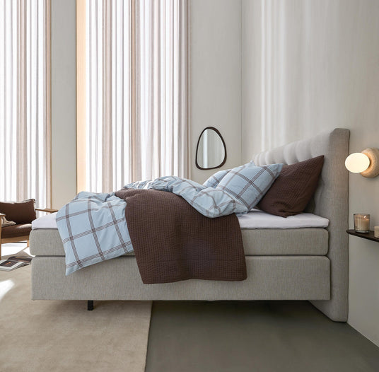 Lyst soveværelse med beige polstret seng, blåternet sengetøj og brun plaid; rund væglampe, lille spejl og lette gardiner i blødt dagslys.