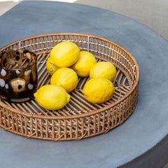 Flettet bakke med gule citroner og et prikket glaslys på et gråt betonbord ved siden af sort vase i dagslys.