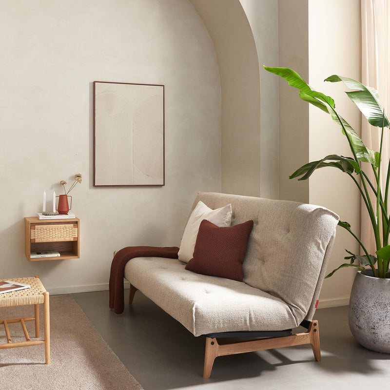 Lyst skandinavisk stue med beige sofa på træben, rustfarvet pude og plaid, stor grøn plante, svævende hylde med vase og lys samt flettet sofabord i neutrale nuancer.