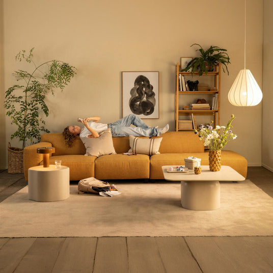 Stue med sennepsgul modulsofa, person ligger med telefon, beige tæppe, runde sideborde, hængende pendellampe, planter og vægbillede.