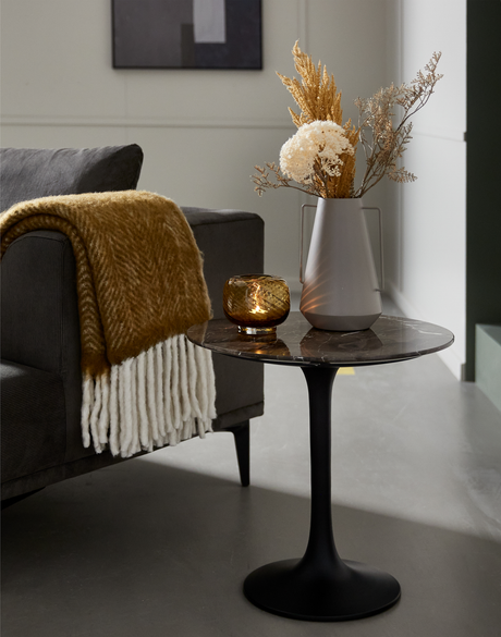 Lille rundt sidebord i mørk marmor med sort trompetfod ved siden af en grå sofa, med ravfarvet lyslygte og grå vase med tørrede blomster samt sennepsgul plaid.