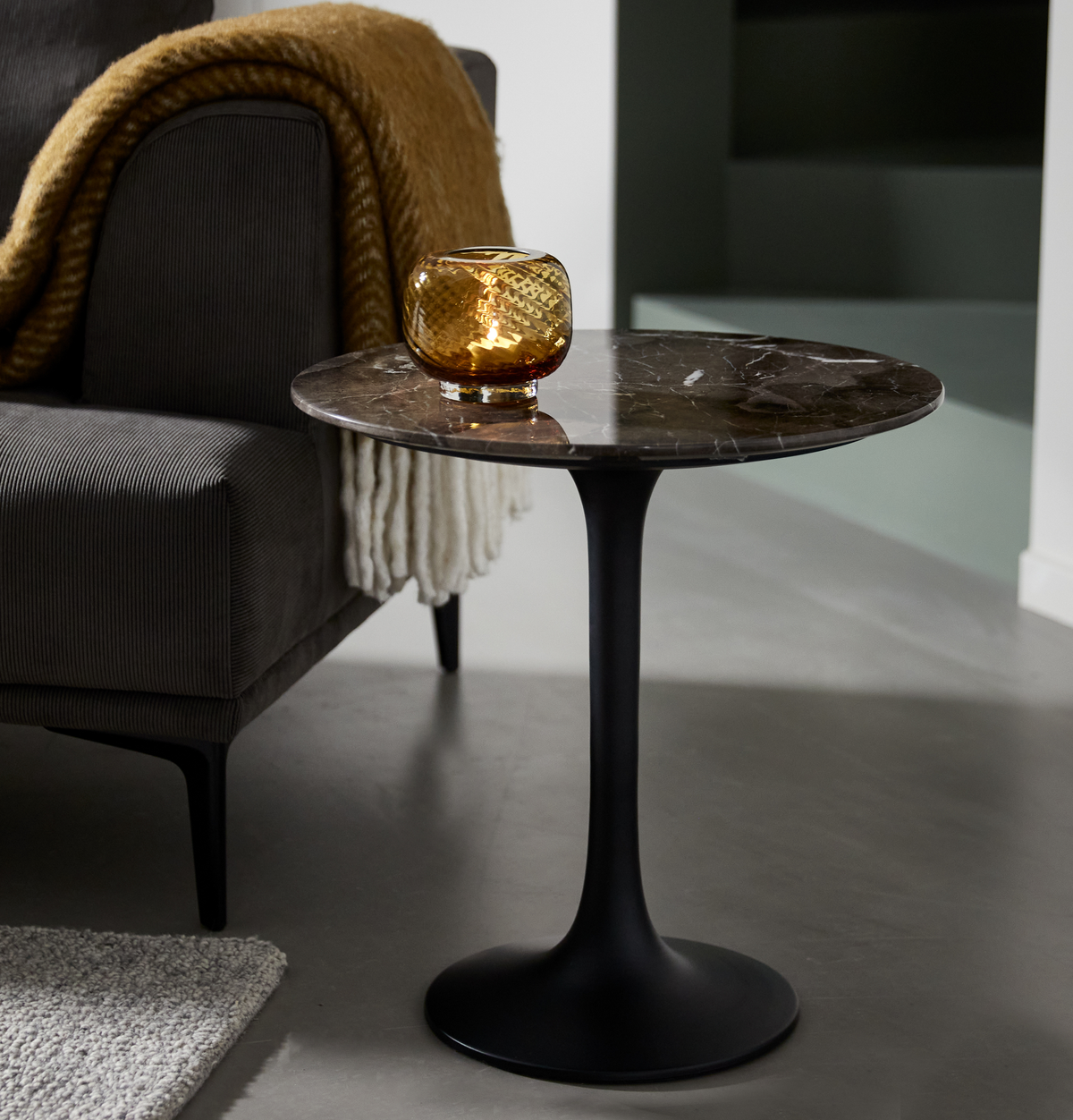 Rundt sidebord i mørk marmor med sort piedestal, ravfarvet lyslygte med tændt lys ovenpå, ved siden af en grå sofa med sennepsgul plaid og hvid frynset tæppe.