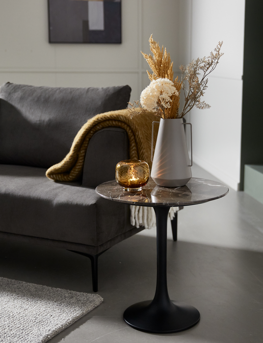 Stuehjørne med grå sofa, sennepsgul plaid og rundt sidebord i mørk marmor med sort fod, vase med tørrede blomster og ravfarvet lyslygte.