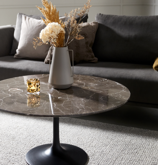 Nærbillede af rundt marmorsofabord med sort fod, vase med tørrede blomster og ravfarvet lyslygte foran en grå sofa i stuen.