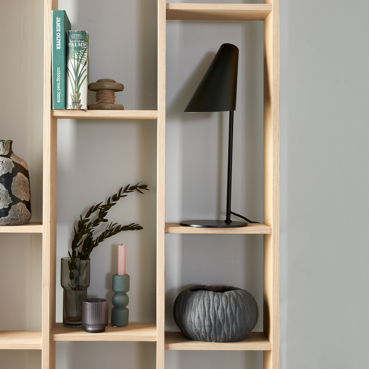 Træreol med sort bordlampe, glasvase med grønne grene, lyserøgt lys i formet lysestage, grå keramikskål og bøger med rygteksten “JAMIE OLIVER” og “PALMS”.