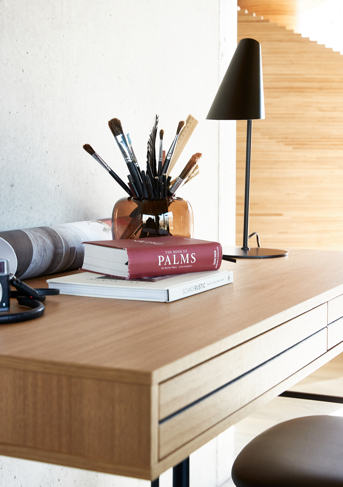Skrivebord i træ med bordlampe, pensler i ravfarvet vase og to bøger, hvor den øverste hedder ”The Book of Palms” og den nederste ”Scandi Rustic”, i et lyst hjemmekontor.
