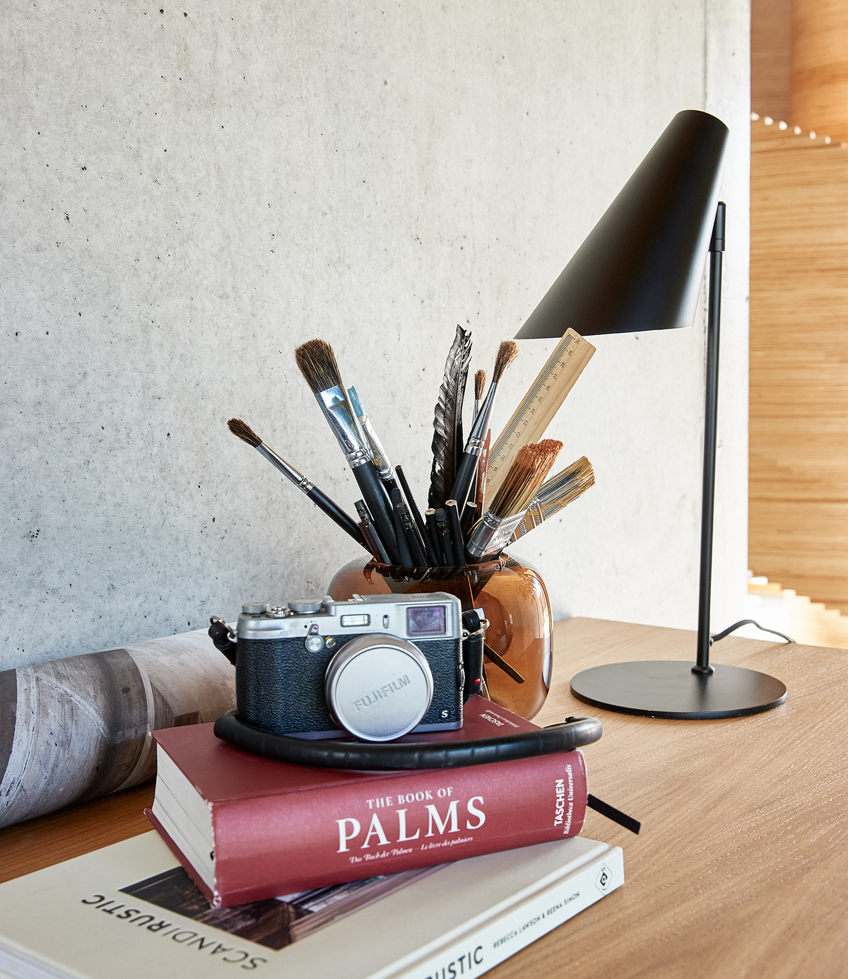 Skrivebord med sort bordlampe, brun glasvase med pensler og lineal, Fujifilm‑kamera oven på en bogstak med The Book of Palms øverst.