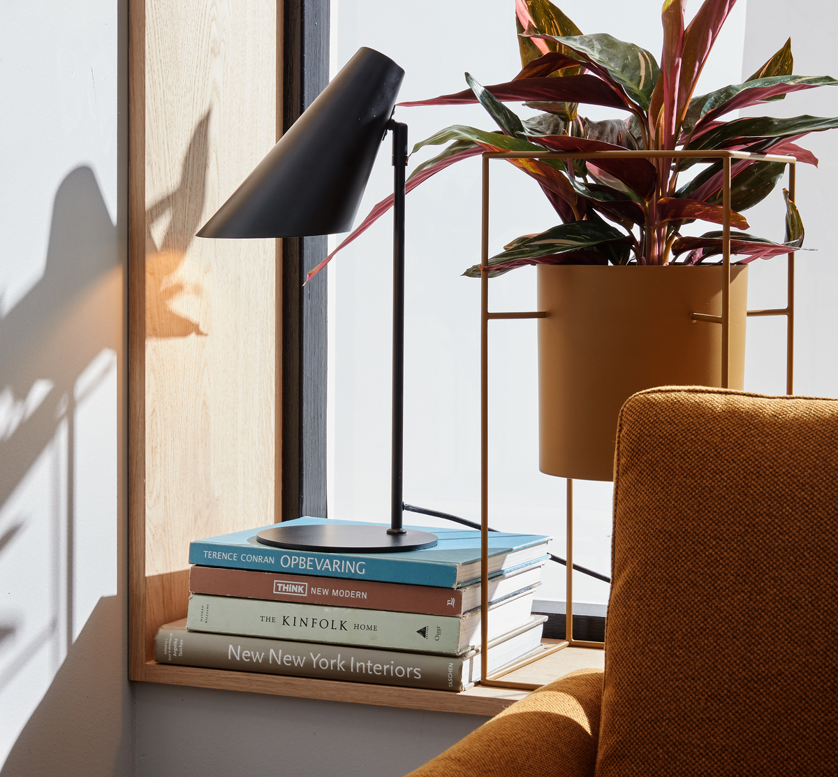 Solbeskinnet vindueskarm med sort bordlampe, en potteplante i sennepsgul stander og en bogstak med titlerne New New York Interiors, The Kinfolk Home, Think New Modern og Terence Conran Opbevaring ved siden af en sennepsgul lænestol.