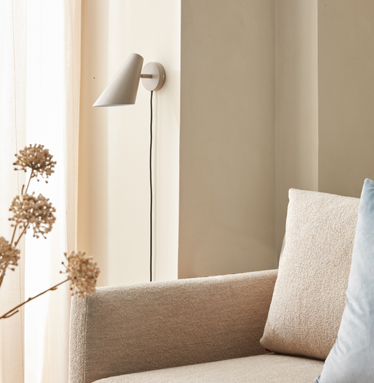 Beige stue med vægmonteret hvid kegleformet væglampe over en beige sofa, blødt dagslys gennem lette gardiner og tørrede blomster; lyseblå pude som accent.