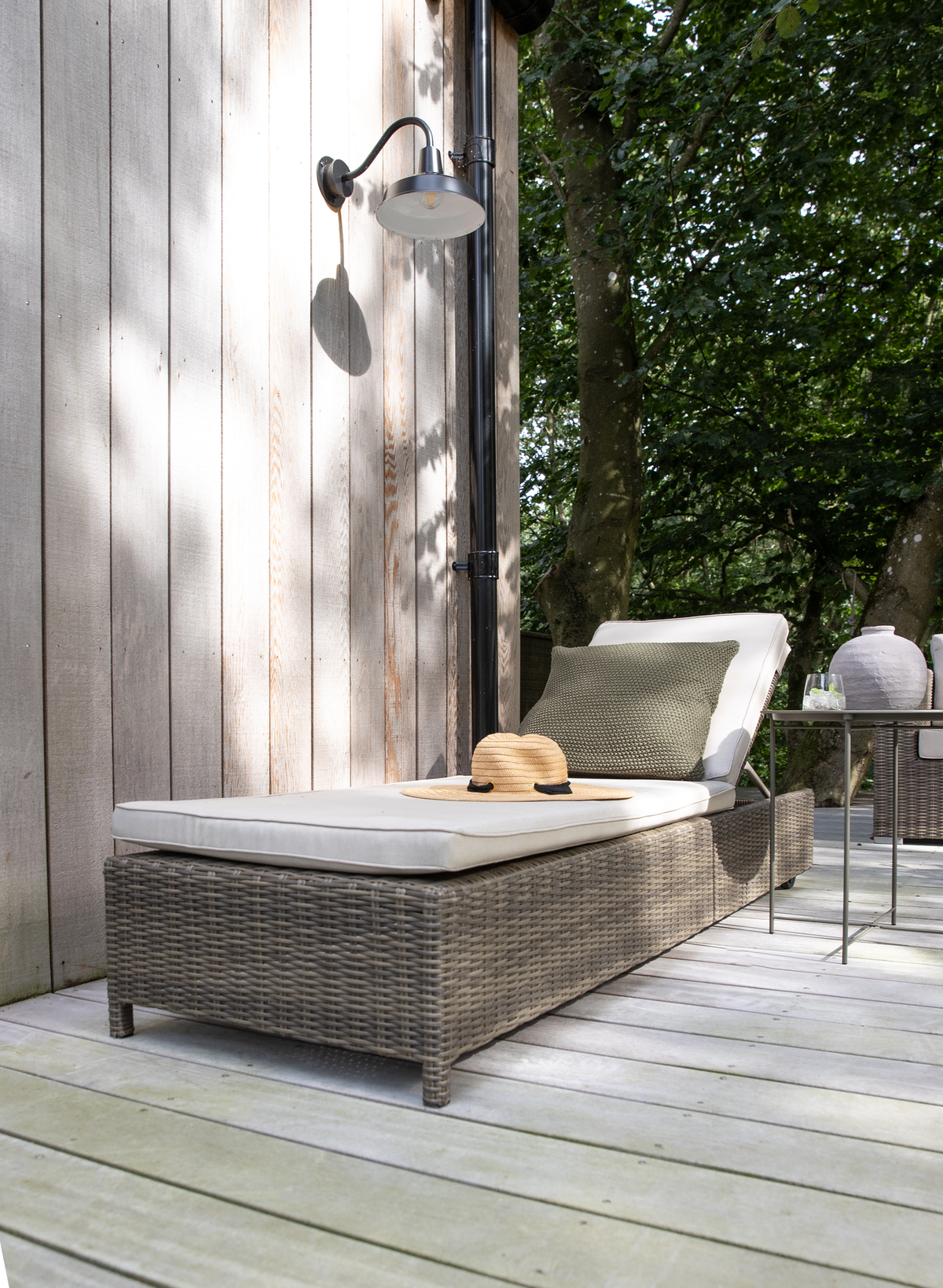 Rattan solseng med lys madras og olivengrøn pude på en træterrasse, med en stråhat ovenpå, vægmonteret udendørslampe og et lille sidebord med glas og keramisk vase i træernes skygge.
