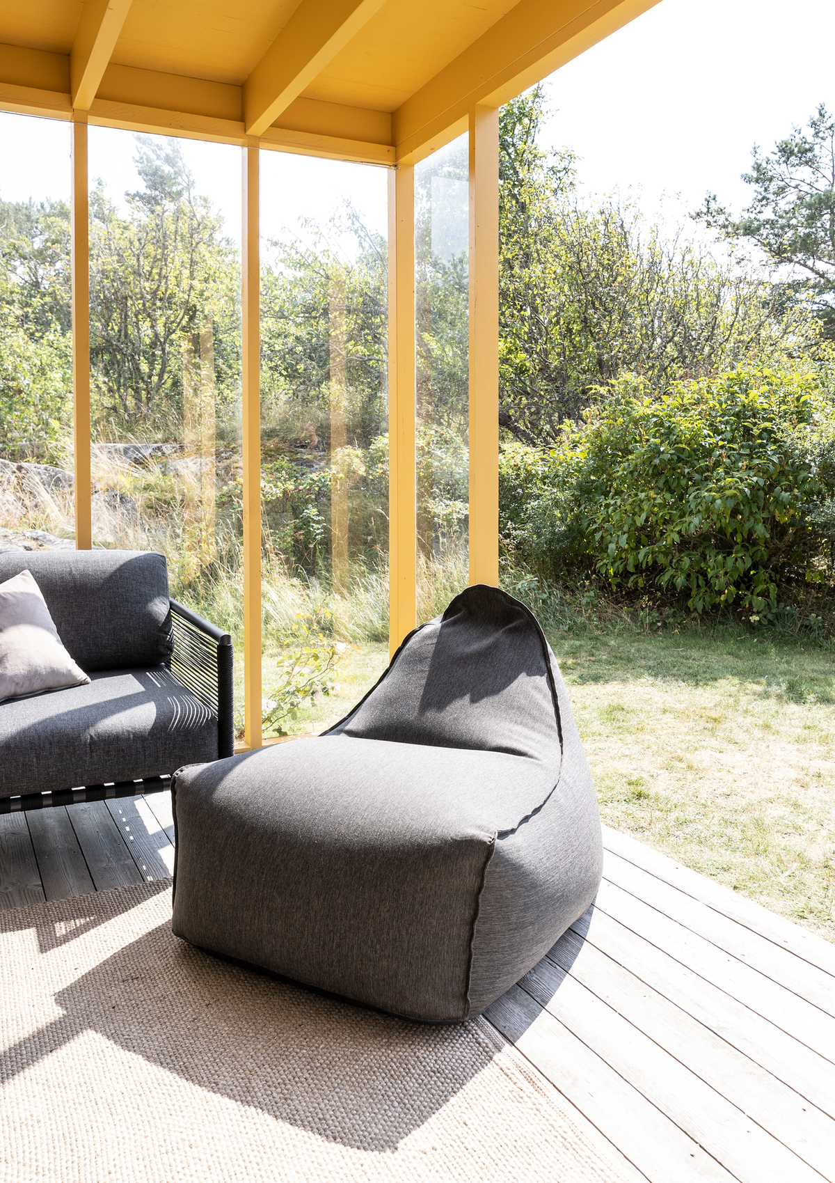 Terrasse med gulmalet veranda, grå sækkestol og grå sofa på trægulv i sollys med grøn beplantning udenfor.